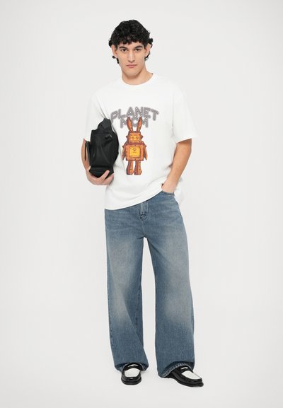 Λευκό t-shirt με εικόνα ενός ρομπότ και το κείμενο "PLANET MCM", συνδυασμένο με ανοιχτό γαλάζιο τζιν παντελόνι με φαρδιά μπατζάκια, μαύρη τσάντα και μαύρα παπούτσια χαμηλής κορυφής.