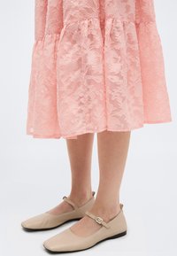 Φλατ παπούτσια Mary Jane σε ανοιχτό μπεζ χρώμα με μικρά λουράκια και αγκράφες, φορεμένα με ροζ φούστα μέσης μήκους με δαντέλα και λουλούδια.