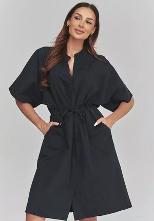 Kobieta o długich brązowych włosach, ubrana w czarną sukienkę do kolan z krótkimi rękawami i wiązaniem w talii, ręce spoczywają w kieszeniach, patrzy na bok.