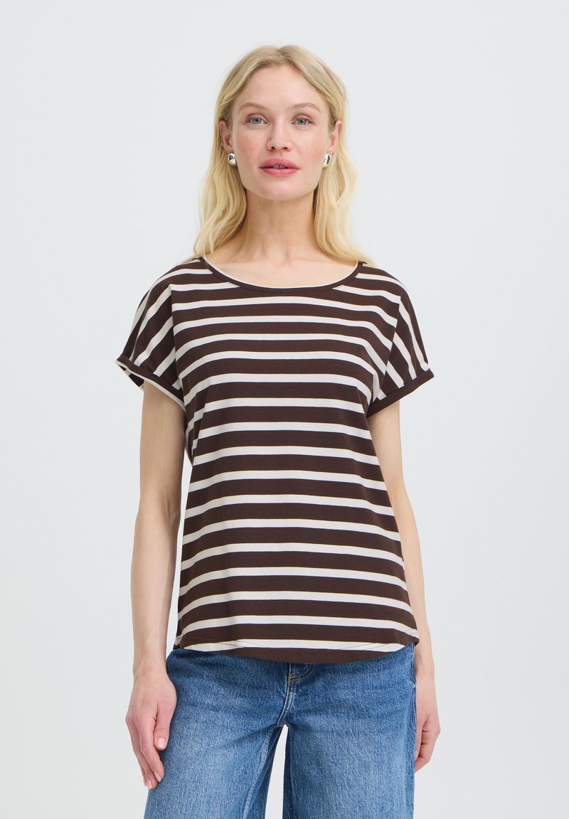 Femme blonde portant des boucles d'oreilles en argent, un t-shirt à manches courtes rayé marron et blanc, et un jean bleu, debout devant un fond clair uni.