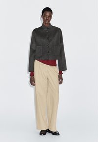 Kobieta w ciemnoszarym zapinanym płaszczu, w czerwono-czarnym pasek topie, beżowych szerokich spodniach i czarnych butach na jednolitym białym tle.