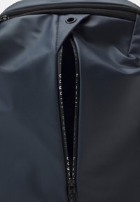 Sinisli vedenpitävä reppu, jossa on vetoketjullinen avaus, sileä rakenne, mustat osat ja näkyvät sisävuoren yksityiskohdat.