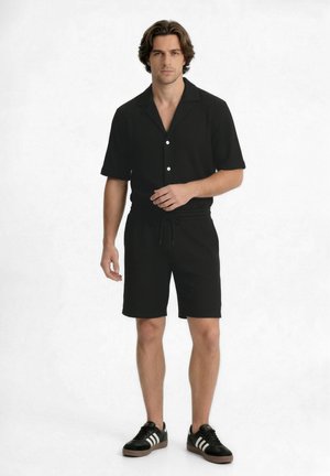 Man standing wearing a black short-sleeve button-up shirt with matching shorts and black sneakers with white stripes on a white background.