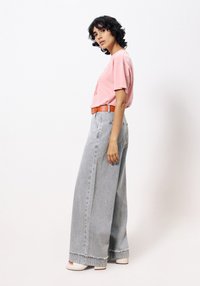 Lichtroze t-shirt met grafische print, gecombineerd met hoge taille, wijdvallende grijze denim jeans met franjes aan de onderkant, en een oranje riem met gesp.