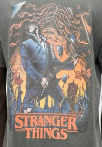 Gray t-shirt featuring an illustration of a guitarist with long hair, surrounded by stylized elements, and "STRANGER THINGS" text in orange.