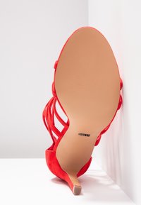 Sandale à talons hauts en suède rouge, dotée d'un design à lanières avec un bout pointu et une semelle lisse couleur beige, positionnée sur fond blanc.