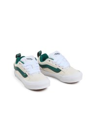 Des chaussures Vans en cuir suédé beige avec des accents verts. Elles possèdent des lacets blancs et une semelle en caoutchouc. Comprend une languette au talon pour un enfilage facile.