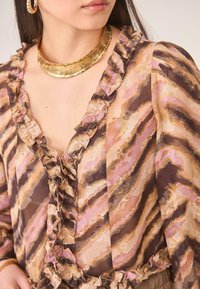 Blusa de estampado marrón y rosa con bordes con volantes, diseño de cuello en V, textura transparente y un collar de declaración dorado como acento.
