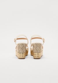 Beige wedge sandals with a woven texture, adjustable ankle straps, and gold-accented platform, featuring a sturdy, patterned heel.