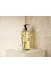Bouteille transparente de shampooing jaune avec pompe noire, partiellement couverte de bulles, placée sur une surface beige texturée contre un arrière-plan neutre.