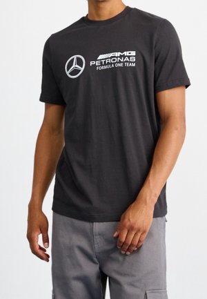 Persoon draagt een zwart AMG Petronas Formula One Team T-shirt en grijze broek, staand met één hand ontspannen langs het lichaam.