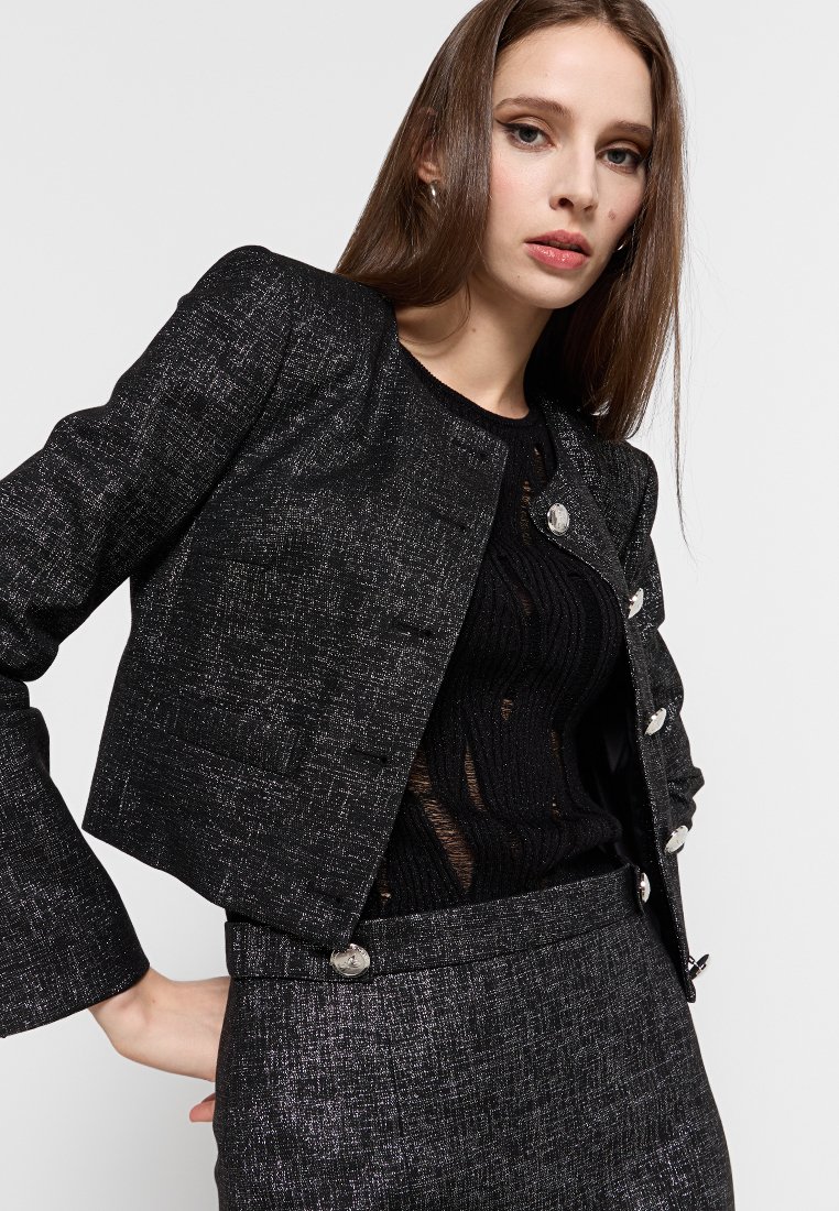 Blazer noir texturé avec un design court, ornée de boutons argentés et de quatre poches, assorti d'un haut noir transparent et d'un pantalon coordonné.