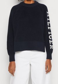 Marineblauer gerippter Strickpullover mit rundem Halsausschnitt und leicht verkürztem Saum. "HILFIGER"-Logo in Weiß auf dem linken Ärmel.