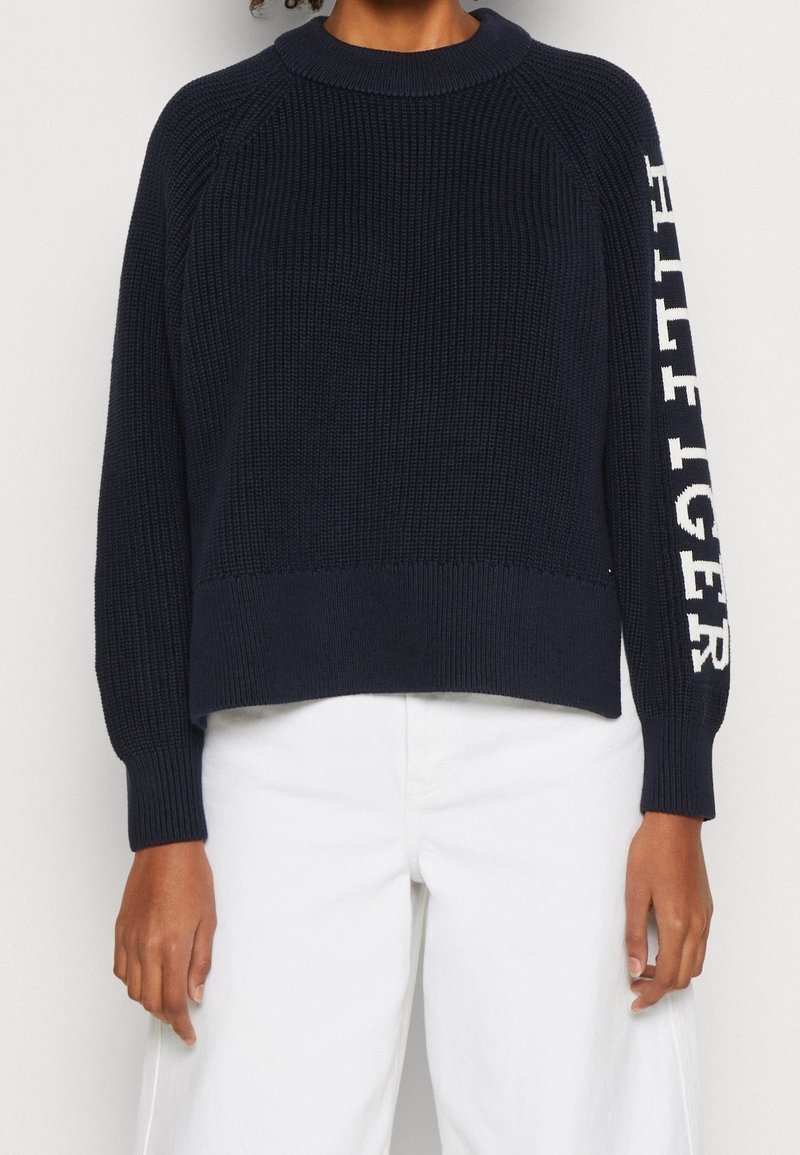 Marineblauer gerippter Strickpullover mit rundem Halsausschnitt und leicht verkürztem Saum. "HILFIGER"-Logo in Weiß auf dem linken Ärmel.