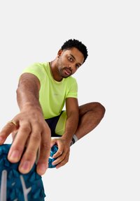 Neono geltonas sportinis marškinėlis su tekstūruotu audiniu, derinamas su tamsiomis šortais. Mėlyni bėgimo batai su ryškiu padu.