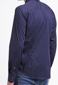 Camicia a maniche lunghe blu navy con polsini bottonati, texture liscia e design aderente. Il retro presenta un yoke e dettagli di cucitura sottili.