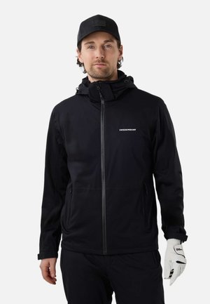 Mand iført en sort Swedemount lynlåstrøje, sort kasket og hvid Wilson golfhandske på højre hånd, stående mod en hvid baggrund.