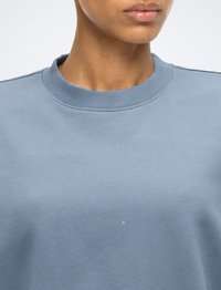 Gros plan d'une personne portant un sweat-shirt bleu clair à col rond, montrant le cou, la ligne de la mâchoire et le haut de la poitrine.