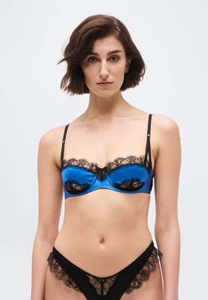 Γυναίκα με μπλε σατέν σουτιέν με μαύρη δαντέλα και αντίστοιχο μαύρο δαντελένιο εσώρουχο, στέκεται μπροστά σε απλό ανοιχτό φόντο.