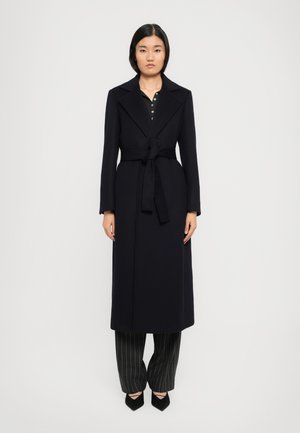 Une femme se tient face à l'avant, portant un long manteau noir ceinturé, un pantalon à rayures noires et des chaussures noires à talons pointus, devant un fond blanc uni.