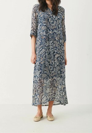 Vrouw draagt een marineblauwe midi-jurk met bloemenpatroon en doorschijnende mouwen, gecombineerd met beige ballerina's, staand tegen een effen witte achtergrond.