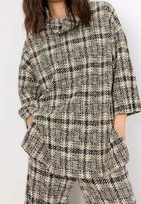 Top de punto a cuadros beige y negro con cuello vuelto, mangas anchas y grandes bolsillos delanteros. Combinado con pantalones a cuadros a juego.