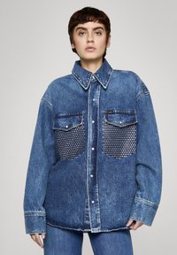 Koszula dżinsowa w ciemnym niebieskim kolorze, z zapinanym na guziki przodem, dużym kołnierzem i dwoma kieszeniami na piersi ozdobionymi srebrnymi ćwiekami.