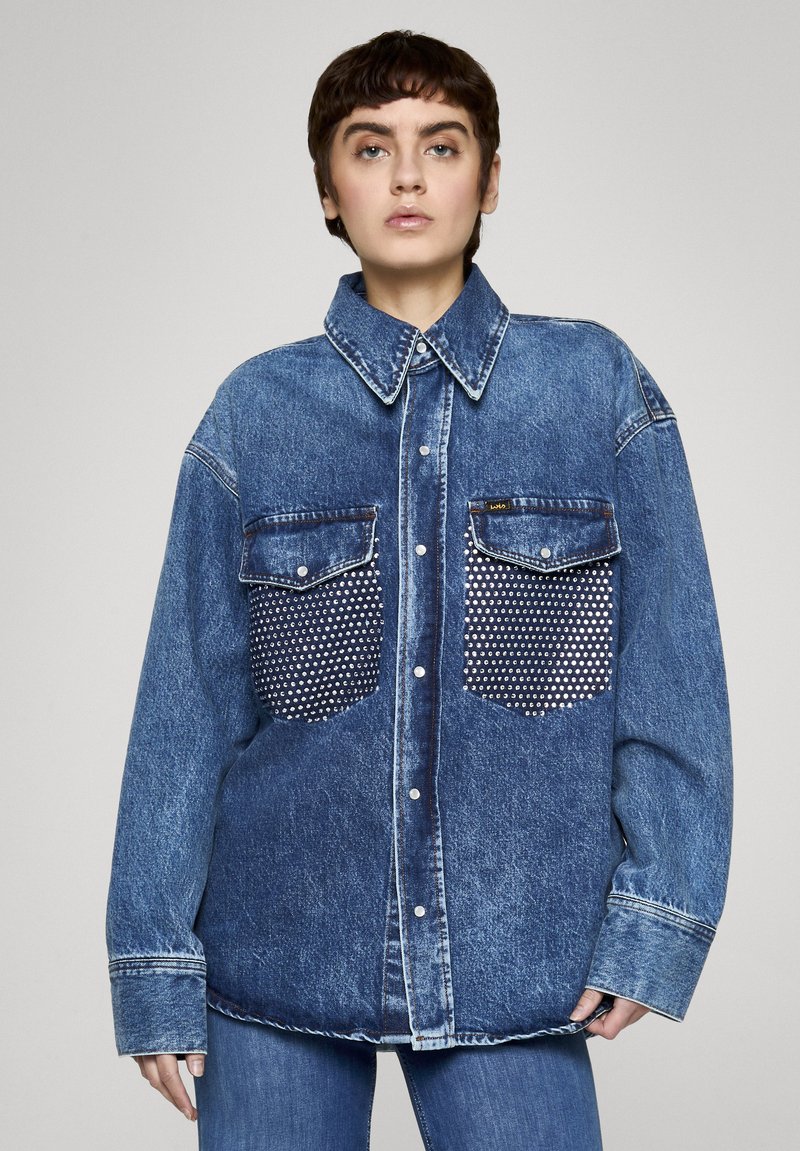 Koszula dżinsowa w ciemnym niebieskim kolorze, z zapinanym na guziki przodem, dużym kołnierzem i dwoma kieszeniami na piersi ozdobionymi srebrnymi ćwiekami.