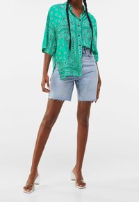 Green paisley-patterned shirt with a loose fit and half sleeves, paired with light denim shorts featuring frayed hems. Clear block-heeled sandals.