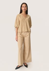 Beige eyelet top with a V-neck, short puff sleeves, and drawstring accents; paired with beige wide-leg trousers and black pointed flats.