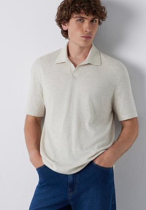 Junger Mann mit lockigem Haar, der ein hellbeiges kurzärmliges Polohemd und blaue Jeans trägt, die Hände in den Taschen, stehend vor einer weißen Wand.