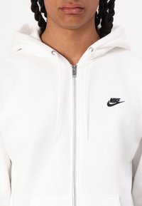 Λευκή φούτερ ζακέτα με φερμουάρ, κατασκευασμένη από μαλακό ύφασμα, με μαύρο λογότυπο Nike στο στήθος και κουκούλα με κορδόνι. Ξεκάθαρα ραψίματα και ασημένιο φερμουάρ.