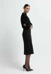 Schwarze Bluse mit langen Ärmeln und strukturiertem Muster, kombiniert mit einem knielangen schwarzen Bleistiftrock. Das Model trägt schwarze spitz zulaufende Absatzschuhe und transparente Strumpfhosen.