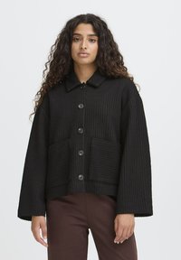 Femme aux longs cheveux bouclés portant une veste noire à boutons avec de grandes poches avant et un pantalon marron, se tenant devant un fond uni.