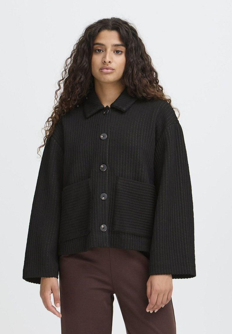 Femme aux longs cheveux bouclés portant une veste noire à boutons avec de grandes poches avant et un pantalon marron, se tenant devant un fond uni.