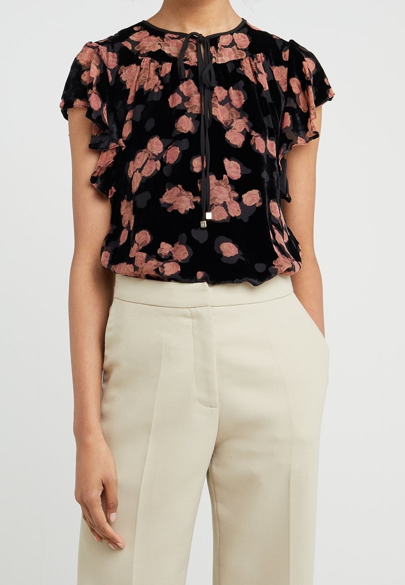 Blouse en velours noir à imprimé floral rose, manches papillon, et col noué, portée avec un pantalon beige clair taille haute.