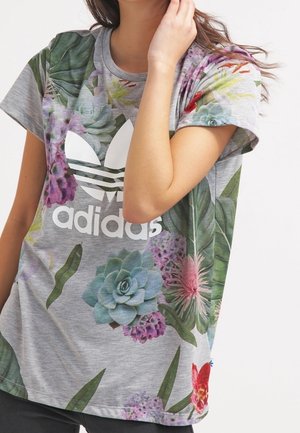 Kobieta ubrana w szarą koszulkę Adidas z dużym białym logo i kolorowymi kwiatowymi oraz sukulentowymi wzorami, pozująca z jedną ręką przy twarzy.