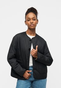 Quiltet sort jakke med rund halsudskæring, frontknapper og påsatte lommer, kombineret med en hvid top og blå jeans. Glat tekstur.