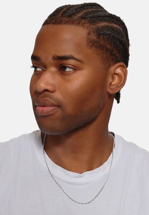 Collier en chaîne argentée porté par une personne avec des cheveux tressés, vêtue d'une chemise blanche, mettant en valeur la texture et les détails du design du collier.