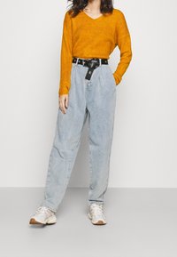 En orange v-ringad tröja kombineras med ljusblå högmidjade jeans. Ett svart bälte med en stor spänne framhäver midjan. Vita sneakers fulländar looken.