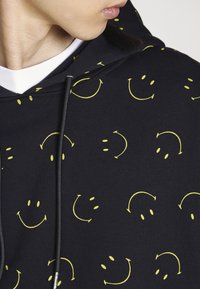 Black hoodie with yellow smiley face pattern, featuring a drawstring hood and soft texture. Close-up of stitching and design detail.
