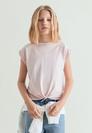 Fată blondă cu păr până la umeri poartă un tricou roz legat și blugi albaștri, cu un hanorac alb legat în jurul taliei.