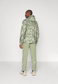 Jachetă ușoară cu model de camuflaj în nuanțe de verde, asortată cu pantaloni corespunzători, având glugă și talie elastică.