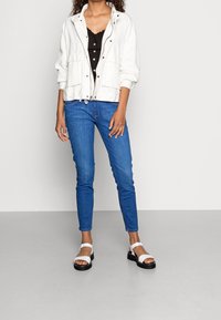 Vit bomulljacka med dragkedja framtill och justerbar dragsko, svart topp med knappar, blå smala jeans och vita sandaler med svarta sulor.