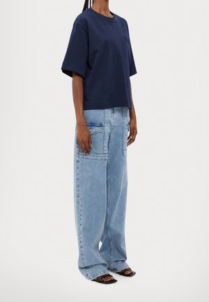 Persona vistiendo una camiseta oversized de color azul marino, jeans de pierna ancha de color azul claro con grandes bolsillos, y sandalias cuadradas de tacón abierto negras.
