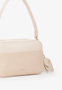 Bolso de mano rectangular beige con un bolsillo frontal de tela texturizada, sección superior de cuero liso, asa corta y etiqueta de equipaje a juego adjunta.