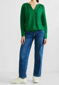 Grön långärmad blus med V-ringning och ruchade axlar, i kombination med vida blå jeans och vita sneakers.