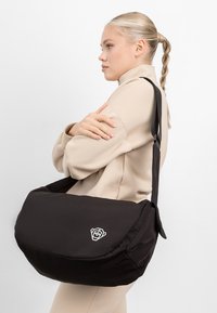 Crna torba za poslane poruke izrađena od mekog materijala, s geometrijskim logom ruže. Dizajnirana s podesivim remenom i strukturalnim oblikom za stabilnost.