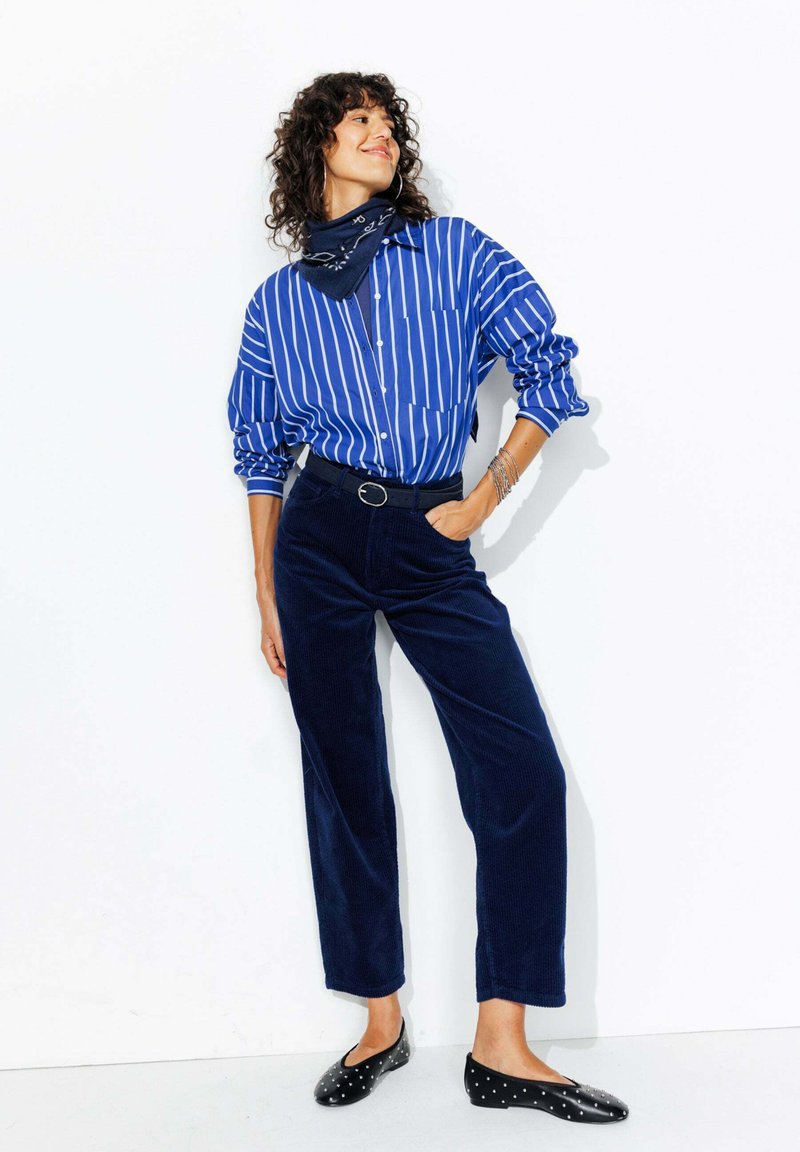 Chemise à rayures bleues et blanches à boutons, pantalon en velours côtelé bleu marine, ballerines noires à pois blancs, et un bandana bleu marine en accent. Bracelets en argent présents.