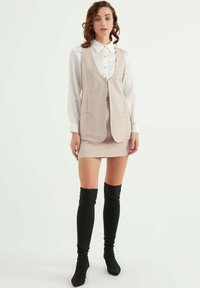 Beige tvådelat set som består av en ärmlös väst med fickor och en figurnära kjol. Kombineras med svarta knähöga stövlar. Texturerad vit skjorta under.